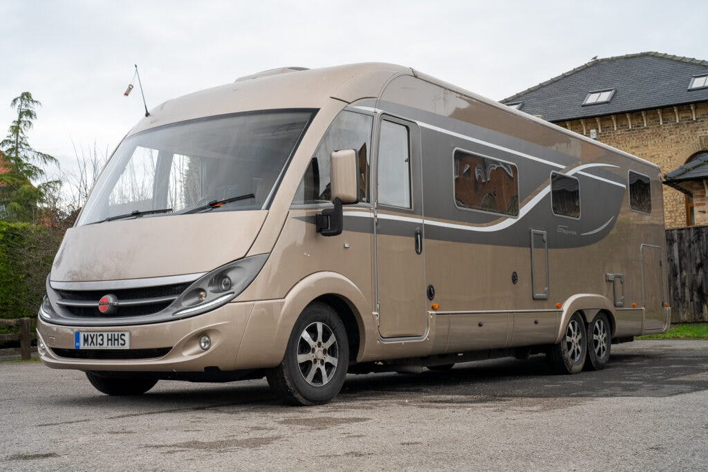 A beige and gray 2013 Burstner Elegance i810 G camper van is parked on a paved area. The vehicle has a streamlined design with multiple windows and is set against a backdrop of a tall brick building with a slate roof. Trees and street signs are also visible in the background.
