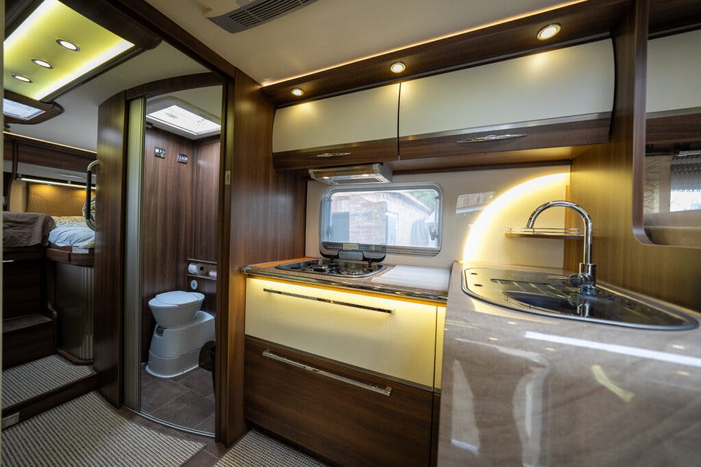 The image shows the interior of a 2013 Burstner Elegance 810 G. It features a kitchen area with a sink, stove, and cabinets, as well as a bathroom with a toilet visible. The space has wooden accents and built-in lighting, with a window above the countertop.