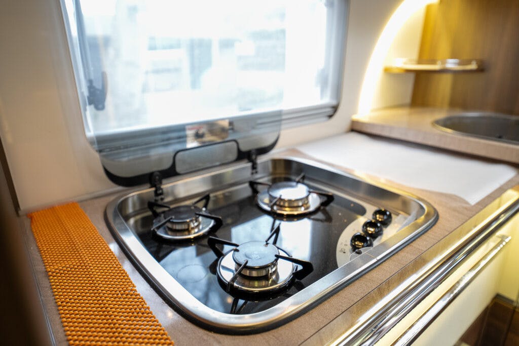 A close-up of a modern kitchen area in the 2013 Burstner Elegance 810 G camper van. It features a built-in two-burner gas stove with black grates and shiny stainless steel surroundings. There is a window above the stove, providing natural light, and a section of the wooden countertop is visible.
