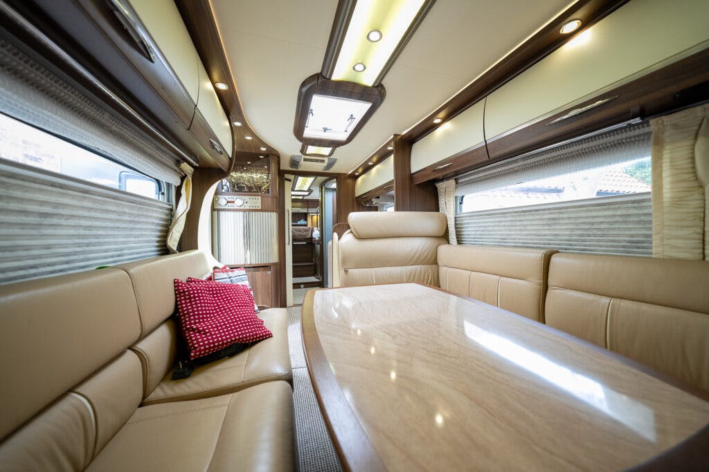 The image showcases the interior of a 2013 Burstner Elegance 810 G luxury motorhome, featuring beige leather seating, a wooden table, and large windows on both sides. The ceiling has recessed lighting and a skylight is visible. Red and black cushions adorn the seating area.