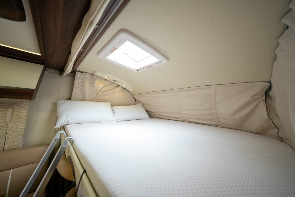 The image shows the interior of a 2013 Burstner Elegance 810 G camper van with a neatly made bed in a loft area. The bed is fitted with white bedding and pillows, and there is an overhead light and small skylight above the bed. A ladder provides access to the bed.