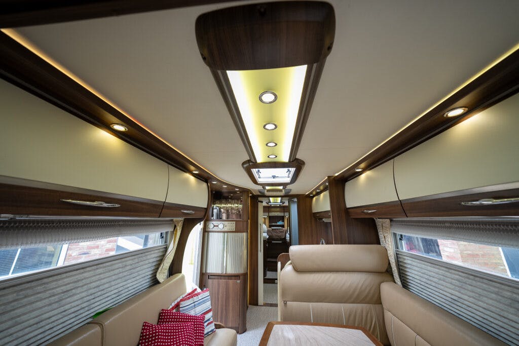 Interior of the 2013 Burstner Elegance 810 G showcasing modern cabinetry and lighting. The ceiling features recessed lights and wooden paneling. On the left is a cushioned seating area with patterned pillows. The rear view shows the corridor leading to additional motorhome sections.