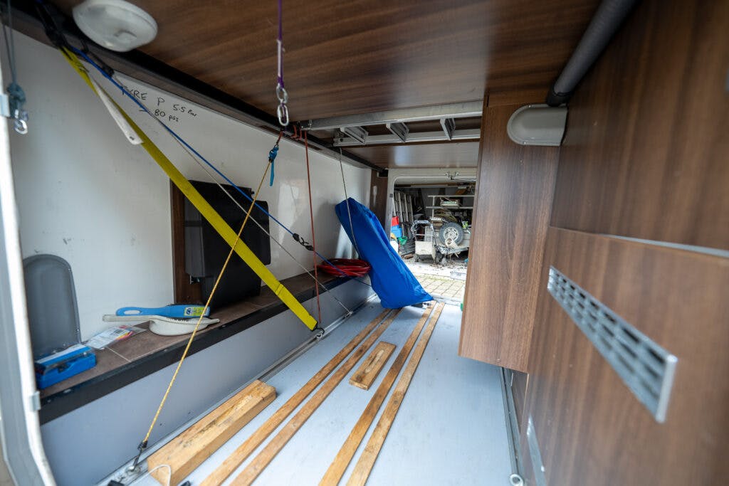 The image depicts the interior of a 2013 Burstner Elegance 810 G camper van or storage area. It contains wood planks, a blue folded item, a yellow strap, and various other items and tools organized within. There is a window providing some natural light and wooden paneling on one wall.