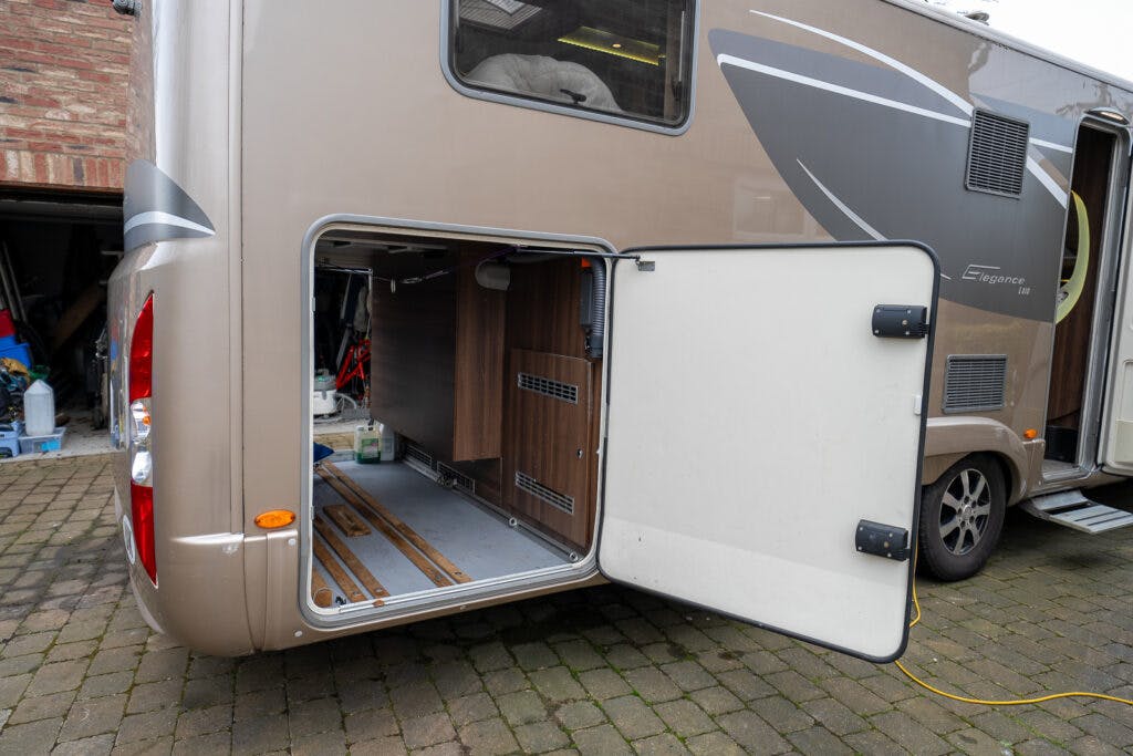 A beige 2013 Burstner Elegance 810 G motorhome is parked on a cobblestone driveway with its side storage compartment open, revealing the interior space containing various items. The compartment door is held open, and a yellow extension cord is plugged into the vehicle.