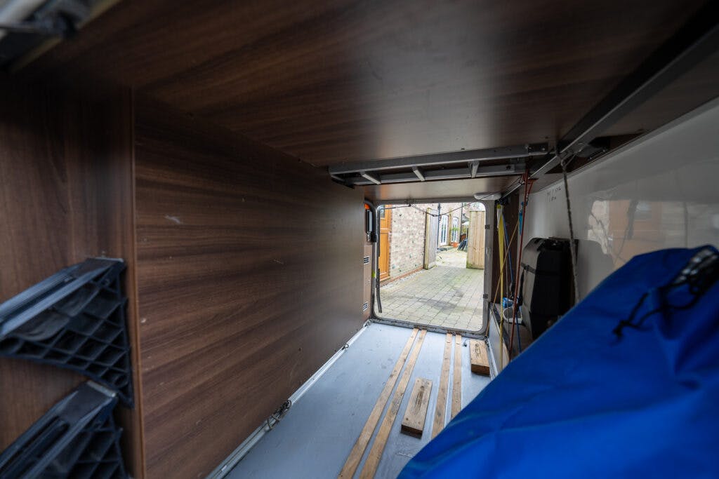 Interior view of the 2013 Burstner Elegance 810 G van cargo area with wooden panels on the sides and metal strips on the floor. The rear door is open, showing a glimpse of the exterior pavement and some buildings. Some cargo appears partially visible near the door.