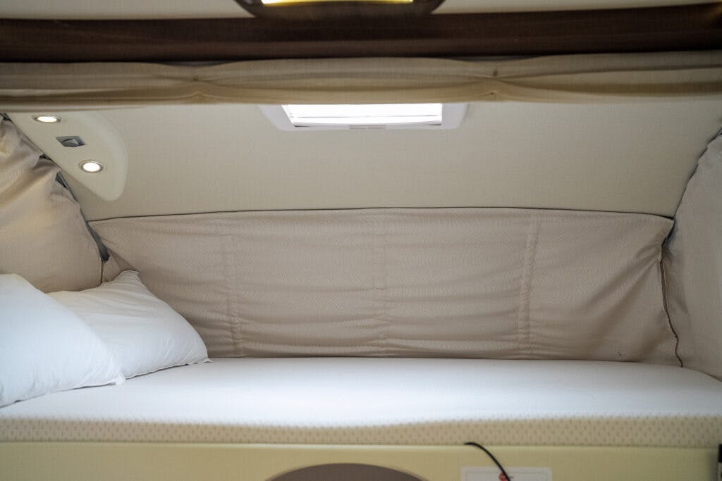 The image shows a small, neatly made bed with white bedding inside a confined space, resembling a sleeping area in the 2013 Burstner Elegance 810 G. Natural light streams through a small window above the bed, and a ceiling-mounted light fixture is visible.