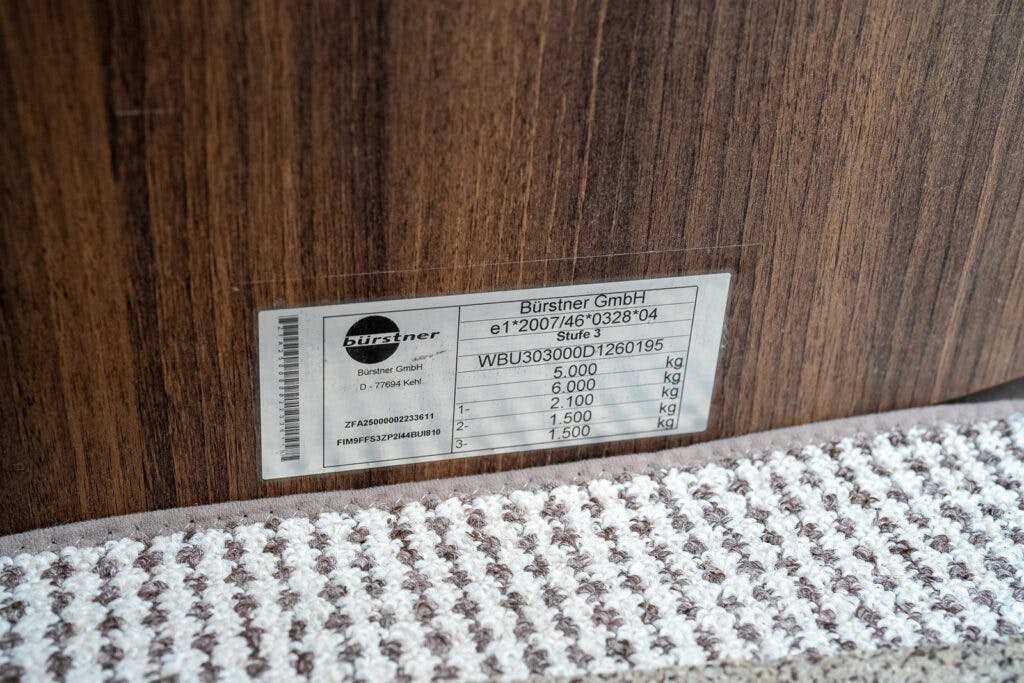 A close-up of a label on a wood-paneled surface with the brand name Bürstner GmbH. The label, from a 2013 Burstner Elegance 810 G, includes specifications and serial numbers, with figures such as "5,000 kg," "6,000 kg," and "1,500 kg" partially visible. A textured carpet edge is seen below the label.