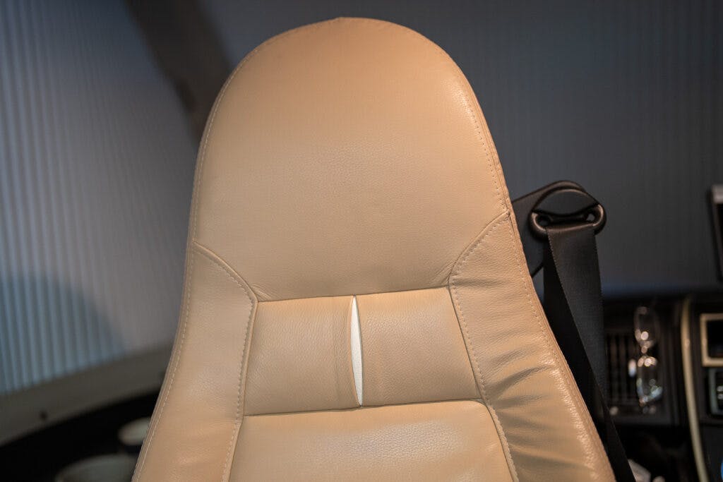 Photo of a light beige leather car seat in a 2013 Burstner Elegance 810 G, featuring detailed stitching and a headrest. The background reveals part of the car's interior, including the seat belt, and the edge of the dashboard in the right corner. The lighting highlights the texture of the leather.
