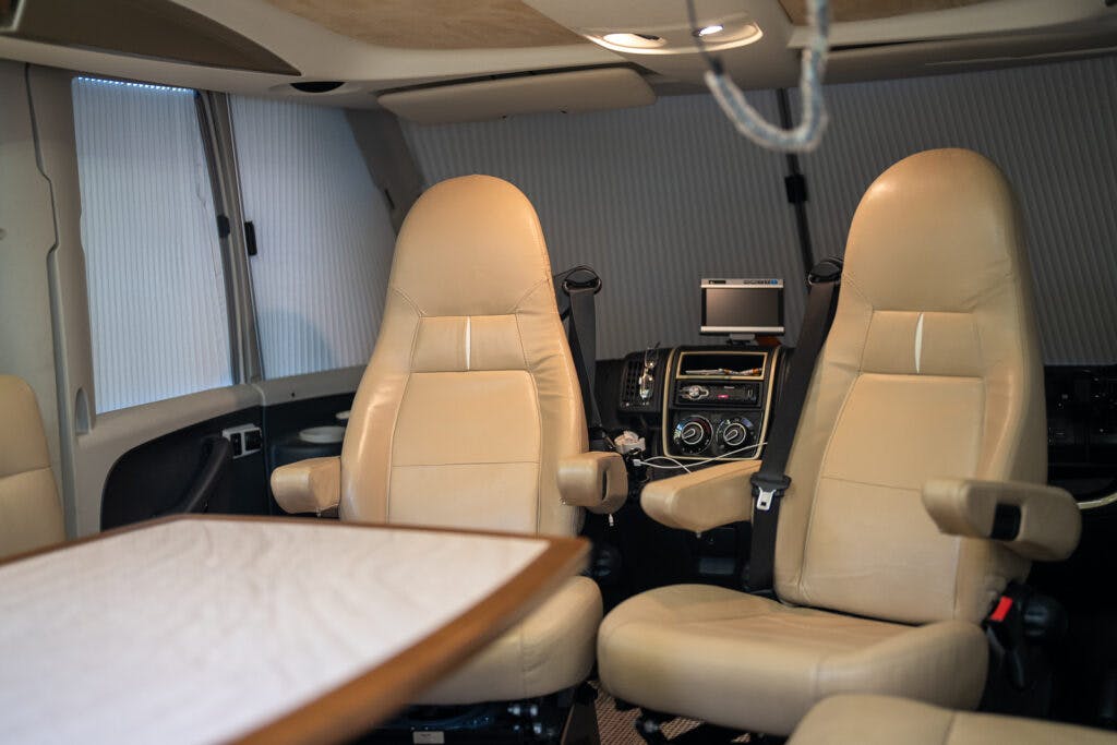 The image shows the interior of a 2013 Burstner Elegance 810 G, likely a recreational vehicle (RV). It features two tan leather seats facing each other, a wooden table, and a compact dashboard equipped with various control panels and a small monitor.