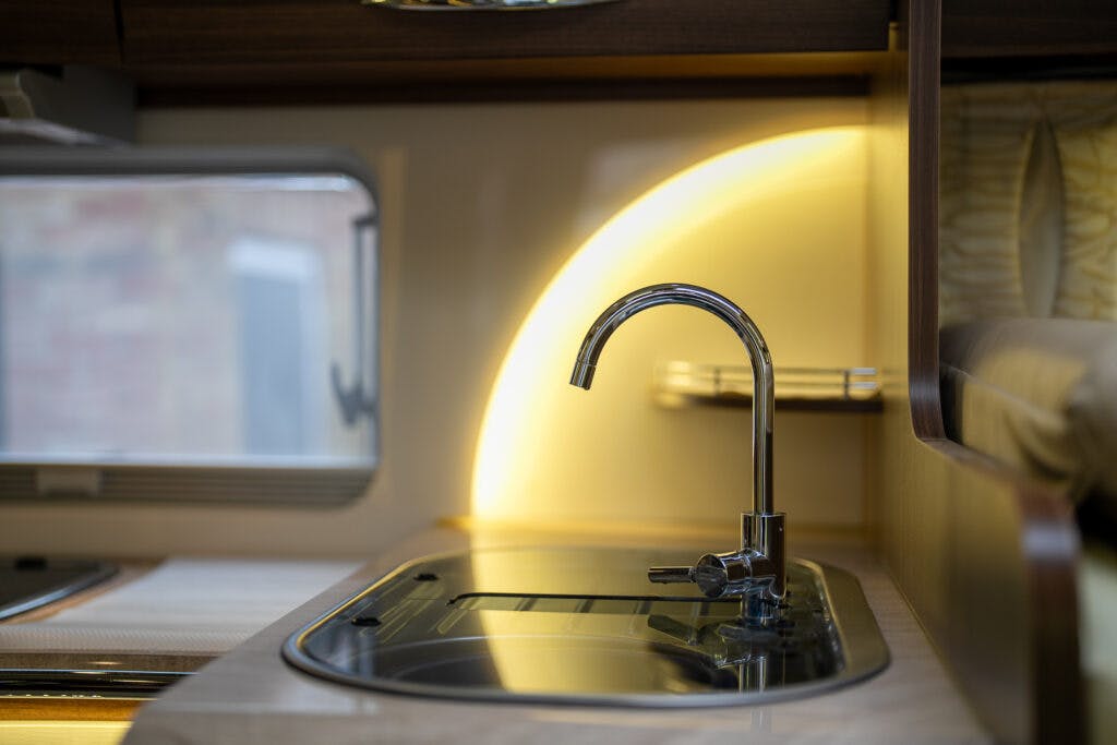 Close-up view of a modern kitchen sink area in a 2013 Burstner Elegance 810 G. The metallic faucet is mounted on a black countertop beside a window. Warm, yellow lighting behind the sink highlights the clean and compact design.