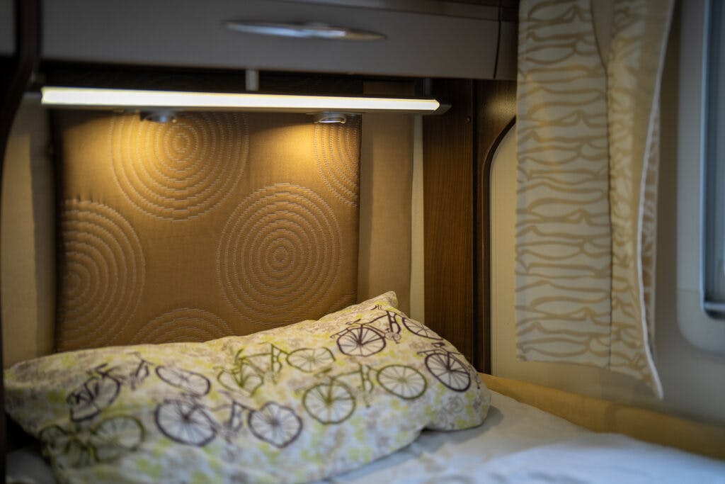A cozy bed in the 2013 Burstner Elegance 810 G RV features a pillow adorned with a bicycle pattern. A soft yellow headboard with circular designs is illuminated by an overhead light, while cream and yellow curtains with a patterned design are partially visible on the right.