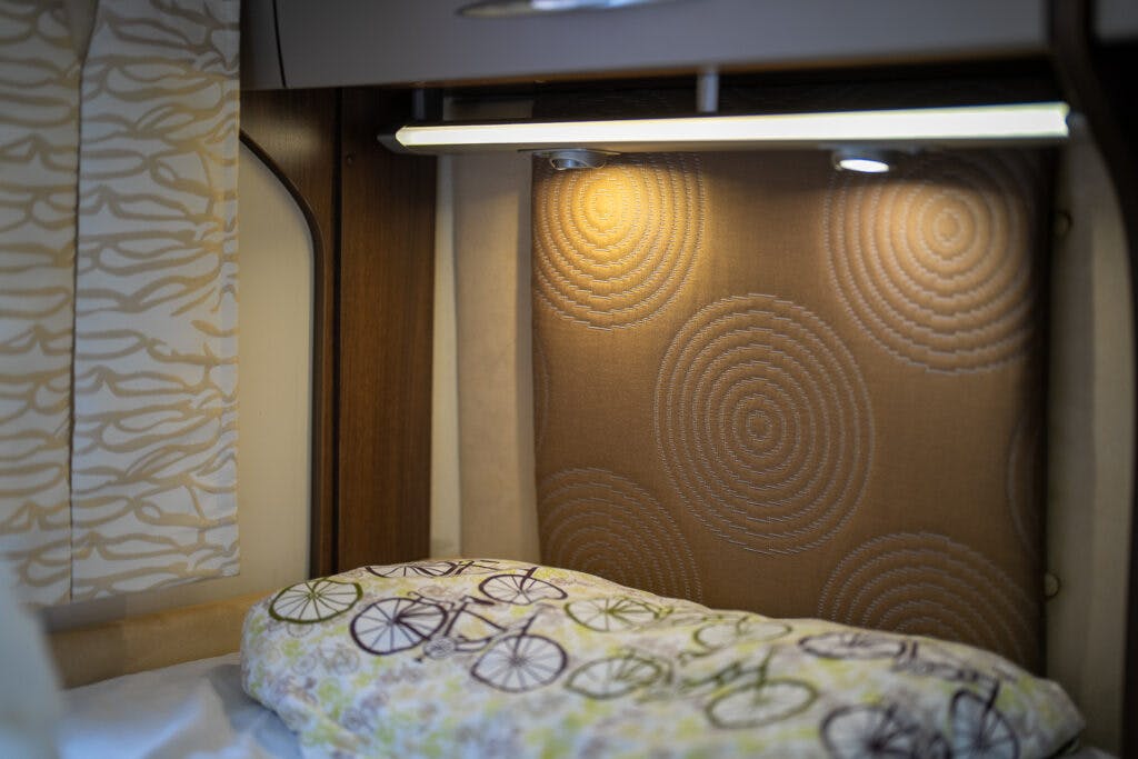 A close-up of a bed in the 2013 Burstner Elegance 810 G, with a light shining above the headboard. The headboard features a textured design with circular patterns. A pillow with a colorful bicycle print is placed at the head of the bed. Curtains with a wavy pattern are partially visible to the side.