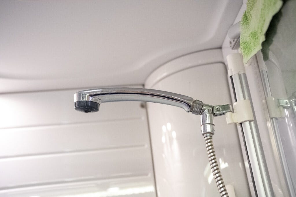 A close-up view of a chrome-plated showerhead attached to a hose in the bathroom of a 2013 Burstner Elegance 810 G. The showerhead is mounted on a white wall, with a green towel hanging in the background.