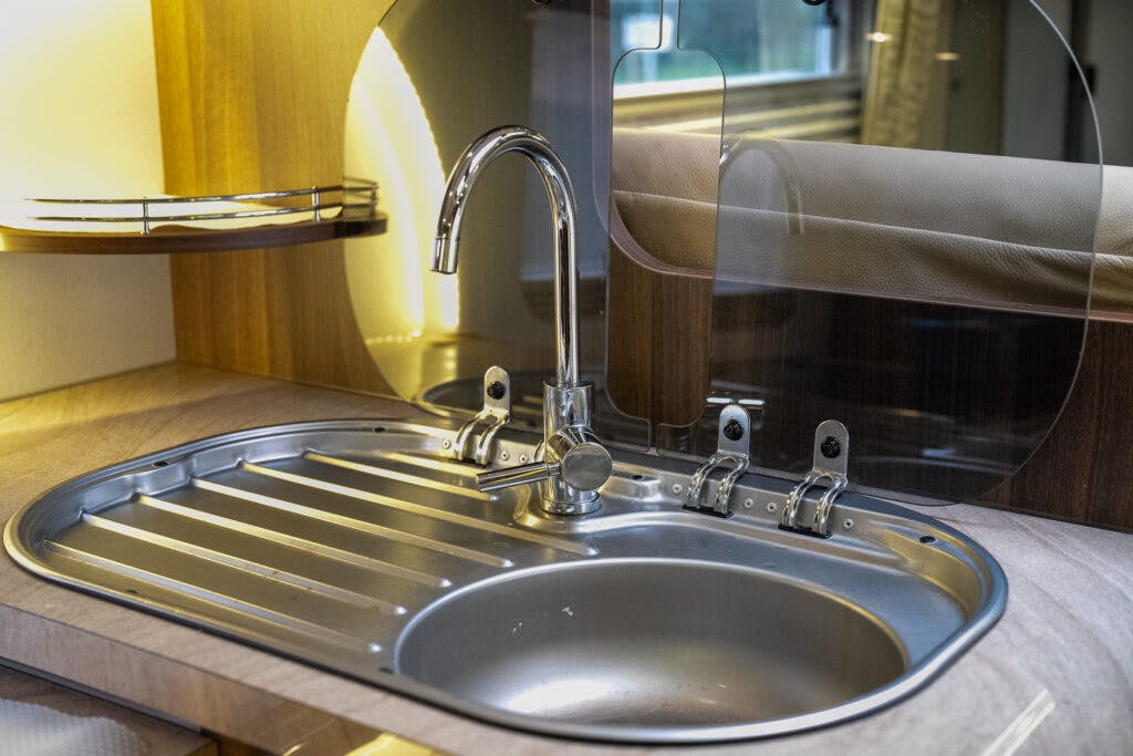 A clean, metal kitchen sink with a curved faucet is situated in a well-lit countertop within the 2013 Burstner Elegance 810 G. Above the sink is a protective glass splash guard. A shelf with rail is visible on the left, and there is a reflection of a window in the splash guard.