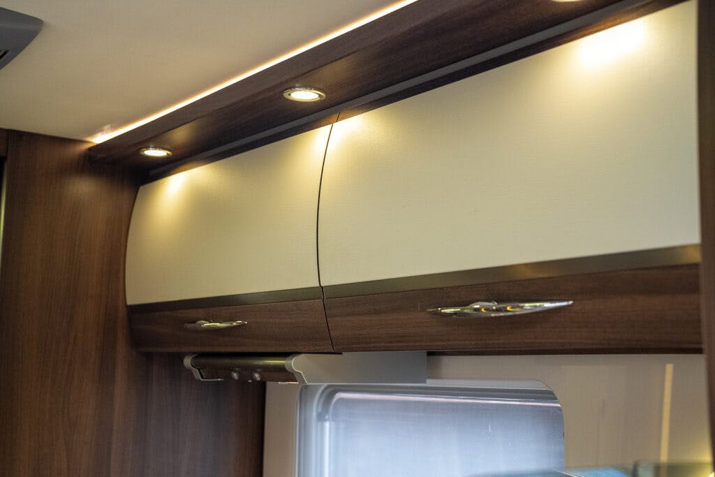 A photo showing the overhead cabinets inside a 2013 Burstner Elegance 810 G. The cabinets have a wood grain finish with white panel accents and metallic handles. Recessed lighting above the cabinets illuminates the area, and a window is partially visible below the cabinets.
