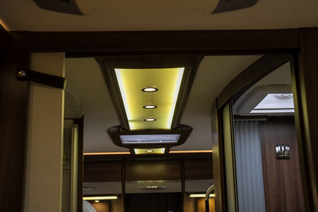Interior view of a 2013 Burstner Elegance 810 G recreational vehicle featuring wooden paneling and ceiling lighting. The ceiling has recessed lights and ambient lighting, with a doorway visible ahead leading to another section of the RV.