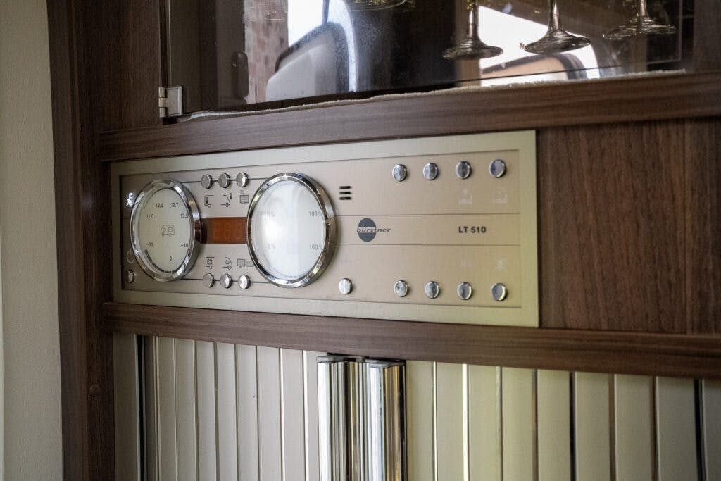A close-up of the control panel on a wooden cabinet in the 2013 Burstner Elegance 810 G, featuring a Schaudt LT 510 display. The panel includes multiple buttons, a digital screen, and two circular dial displays. Glassware is stored in the shelf above. Vertical blinds partially cover the lower section.