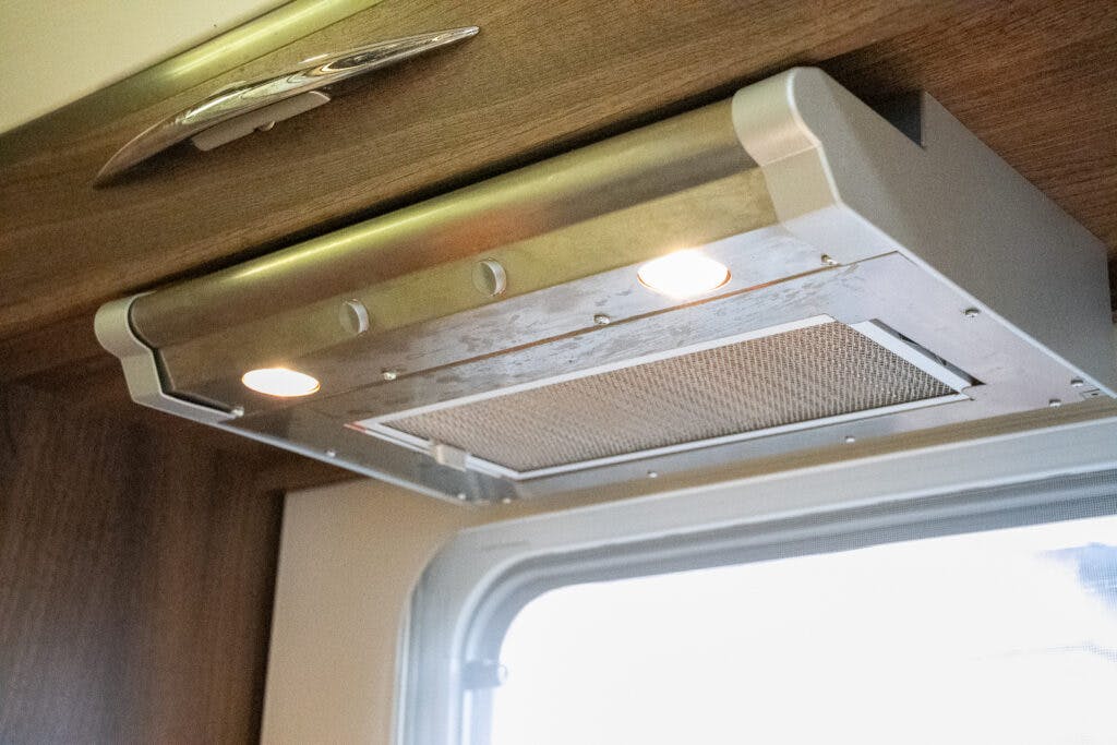A close-up view of a modern under-cabinet range hood with stainless steel finish, illuminated by two small lights. The hood is installed beneath wooden cabinetry, reminiscent of the sleek design found in a 2013 Burstner Elegance 810 G. A window is visible in the background.