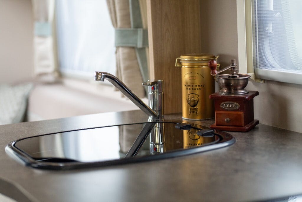 A modern kitchen countertop inside a 2019 Elddis Autoquest 196 Signature Edition camper features a sleek black sink with a chrome faucet. Behind it, a yellow tin labeled "traditional tea bags" and a vintage coffee grinder with a label reading "café" sit against a window with light curtains.