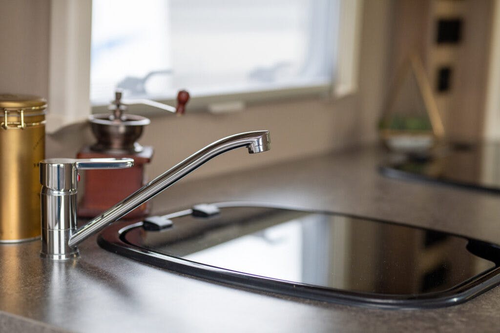 A kitchen sink with a modern, curved faucet is shown with a black covering over the basin. The grey countertop holds a gold canister and a coffee grinder beside the sink. In the background, through the window, you can spot part of a 2019 Elddis Autoquest 196 Signature Edition motorhome parked outside.