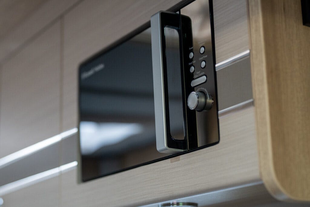A close-up image of a modern microwave oven installed in a wooden kitchen cabinet. The microwave has a digital display, control buttons, and a metallic handle. This sleek setup is reminiscent of the high-end fixtures found in the 2019 Elddis Autoquest 196 Signature Edition motorhome, with the wood grain and reflective surfaces clearly visible.