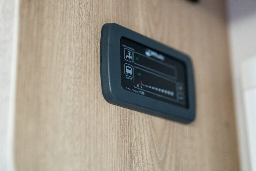 A close-up photo of a control panel mounted on a wooden surface inside the 2019 Elddis Autoquest 196 Signature Edition. The panel features a small screen and several buttons or indicators, with the background softly out of focus.