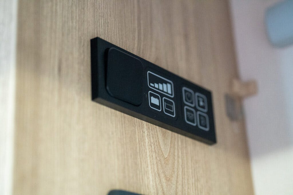 A close-up image of a control panel mounted on a wooden surface in the 2019 Elddis Autoquest 196 Signature Edition. The panel features several buttons and icons, including a battery-level indicator and other various function symbols. The background showcases a light-colored wooden texture.