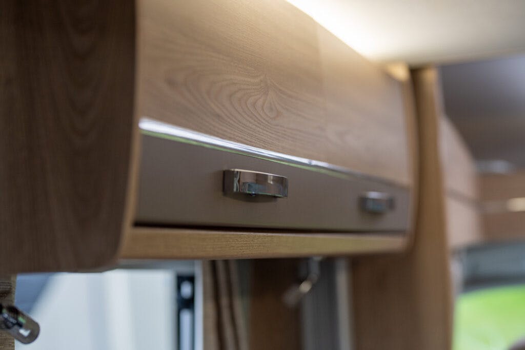 A close-up view of a wooden cabinet with two metal handles in a well-lit room, reminiscent of the sleek design in the 2019 Elddis Autoquest 196 Signature Edition. The cabinet has a modern design and smooth finish, with a window featuring drawn beige curtains partially visible in the background.
