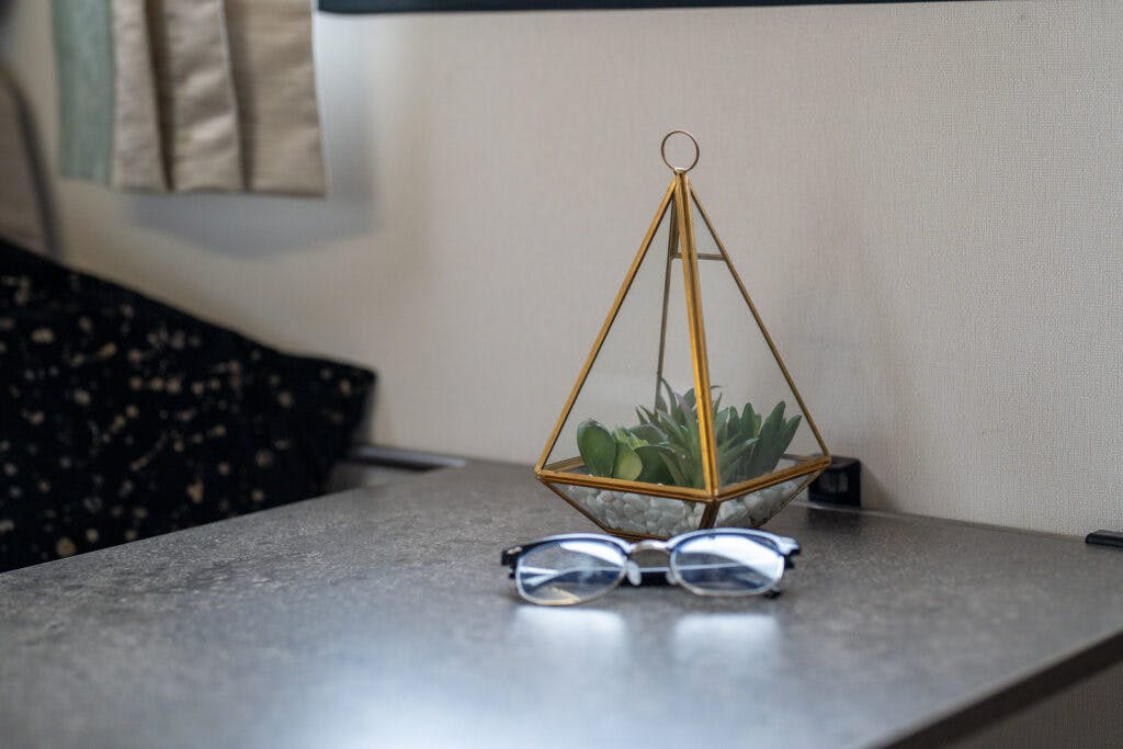 A pair of reading glasses rests on a gray table. Behind the glasses, there is a small succulent plant in a geometric, gold-colored glass terrarium. A beige curtain and a black patterned fabric reminiscent of the 2019 Elddis Autoquest 196 Signature Edition's interior are in the background.