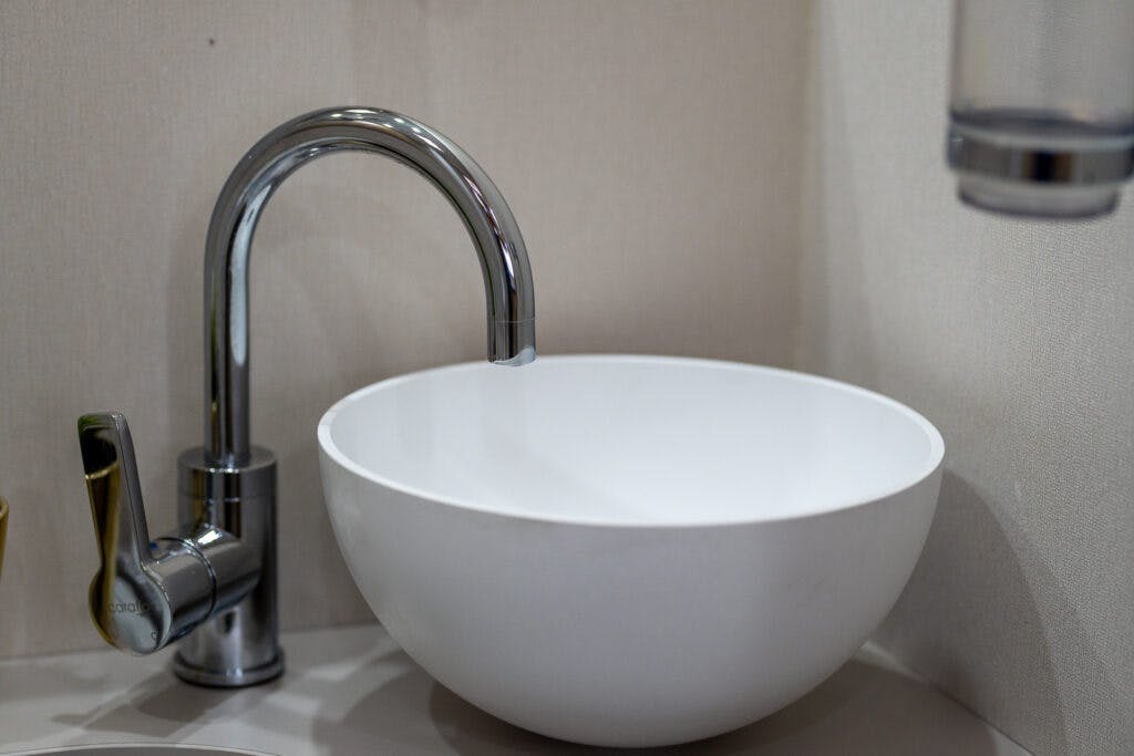 A modern bathroom sink in the 2019 Elddis Autoquest 196 Signature Edition features a white, round basin and a tall, chrome faucet with a single lever handle. The faucet and basin are set against a plain beige background, accompanied by a partial view of a wall-mounted soap dispenser on the right.