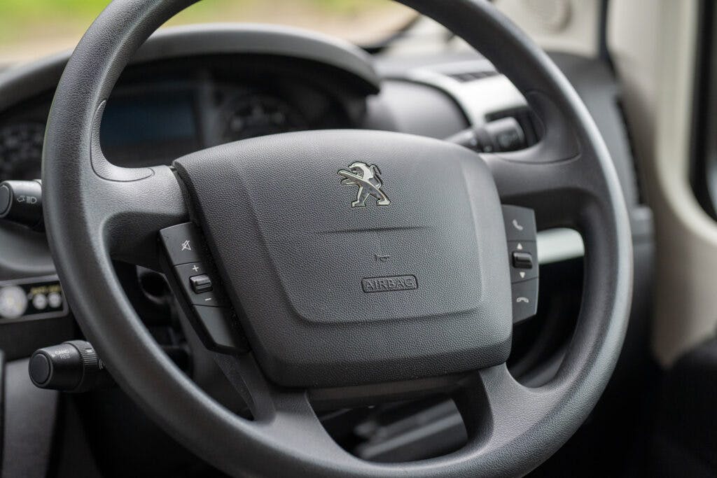 Close-up view of a car steering wheel with the Peugeot logo in the center, featured in the 2019 Elddis Autoquest 196 Signature Edition. The wheel has control buttons for audio and other functions on either side. In the background, the dashboard and part of the air vent are visible.