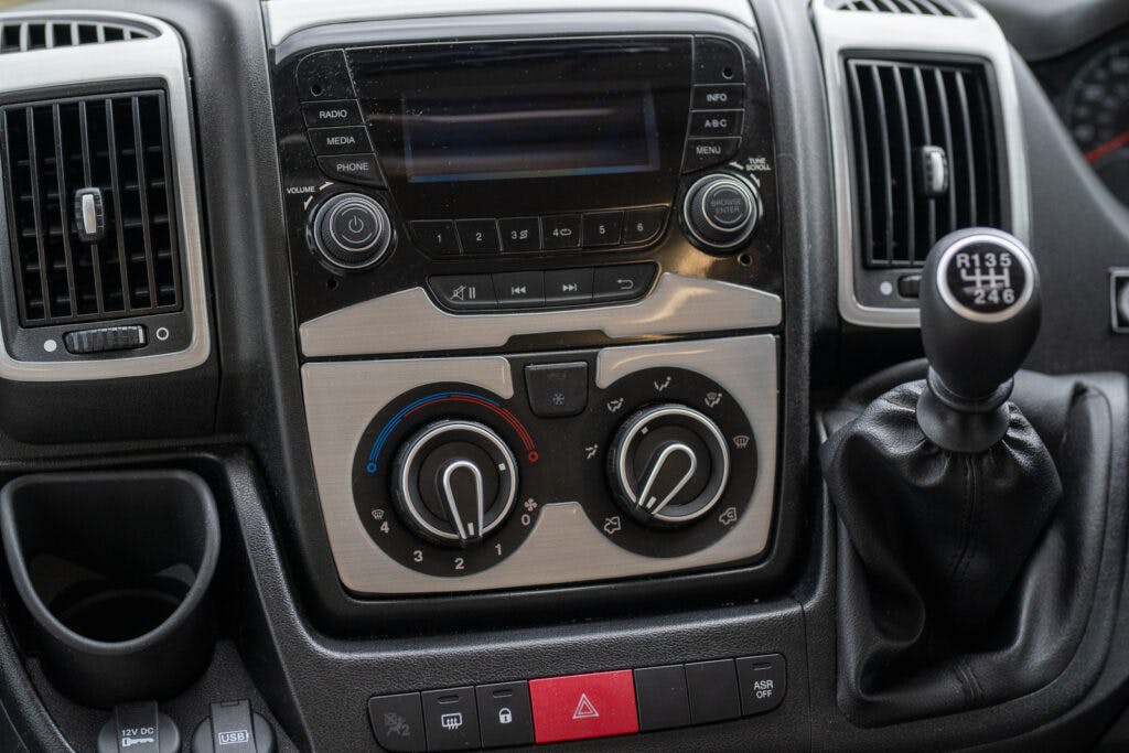 Close-up view of the 2019 Elddis Autoquest 196 Signature Edition's dashboard, featuring a multimedia system with a screen and buttons, two air vents on either side, and several dials and controls for climate settings below. A gear shift lever and various buttons, including hazard and other controls, are also visible.