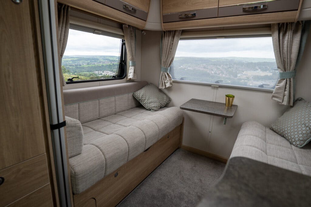 The interior of the 2019 Elddis Autoquest 196 Signature Edition RV features two facing beige cushioned benches with green throw pillows, a small wooden table between them, and large windows with tan curtains offering a view of greenery outside. The space is well-lit, with light wood cabinets overhead.