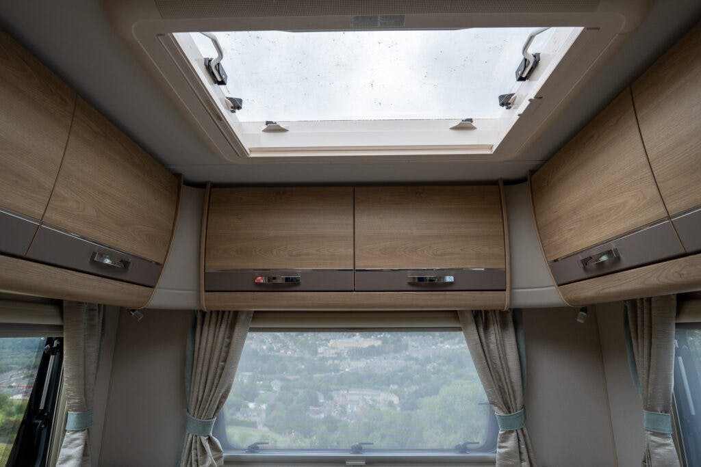 Interior view of the 2019 Elddis Autoquest 196 Signature Edition campervan featuring wooden overhead storage cabinets, grey curtains partially drawn over a large window, and an open skylight window in the ceiling. The window provides a glimpse of a landscape with trees and hills.