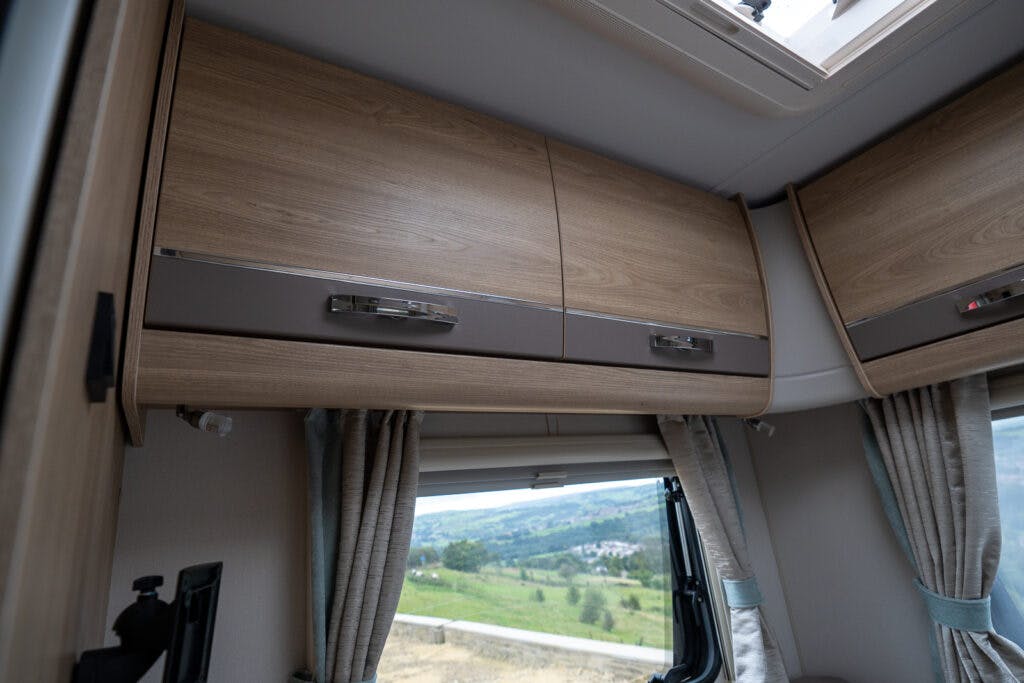 Interior view of a 2019 Elddis Autoquest 196 Signature Edition RV showing overhead wood-paneled storage cabinets with metallic handles. A window with light gray curtains offers a view of a landscape with hills and greenery outside.