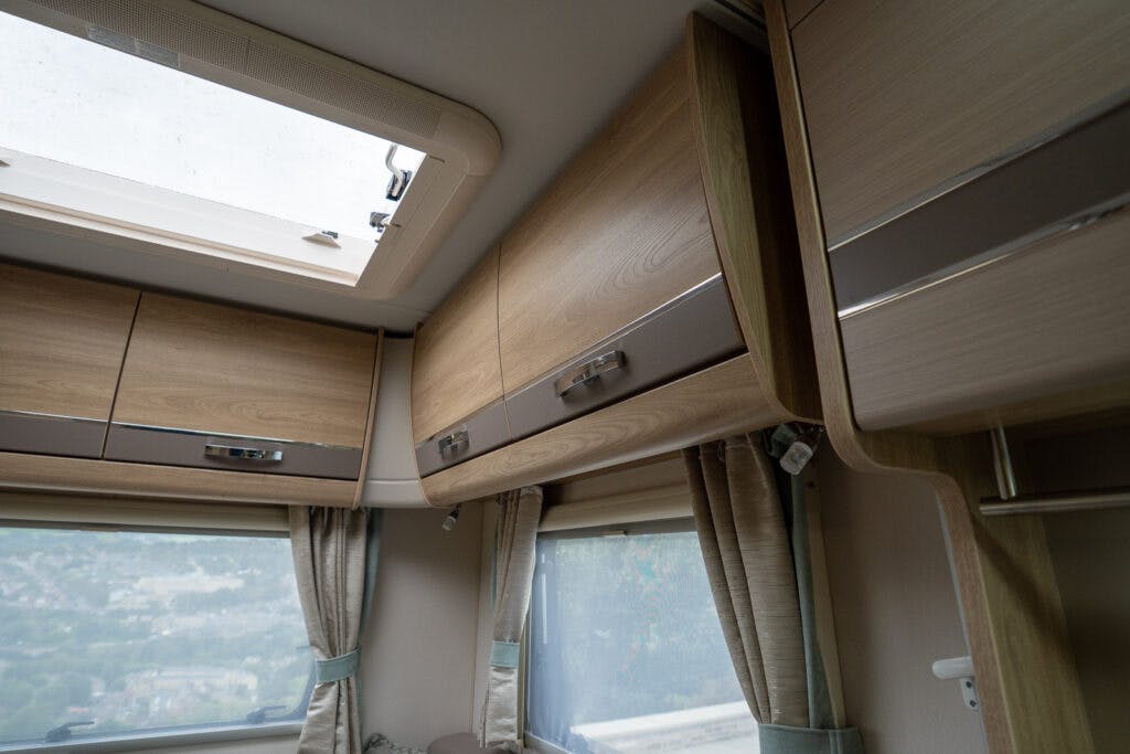 Interior of the 2019 Elddis Autoquest 196 Signature Edition RV showing wooden overhead cabinets with chrome handles, beige curtains on large windows, and a ceiling vent providing natural light.