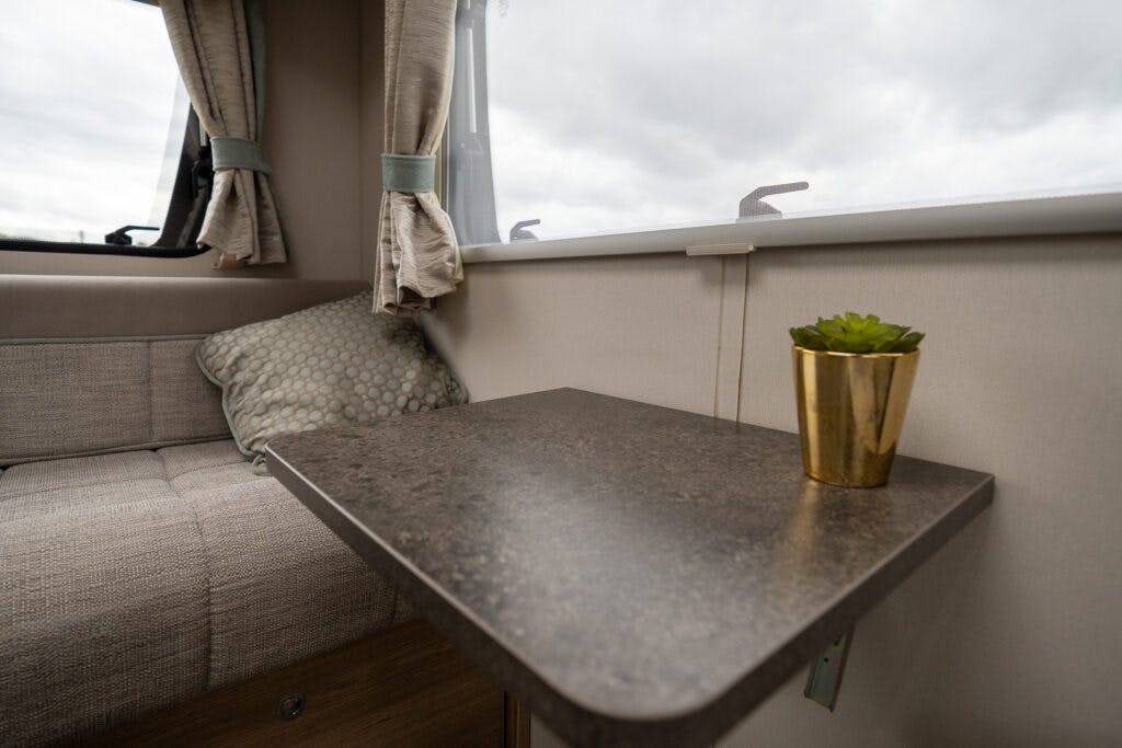 The 2019 Elddis Autoquest 196 Signature Edition boasts a small, modern RV interior featuring a corner with a cushioned bench seat, a window with beige curtains, a polka-dot pillow, and a grey table attached to the wall. Adding charm is a small green succulent plant in a gold-colored pot.