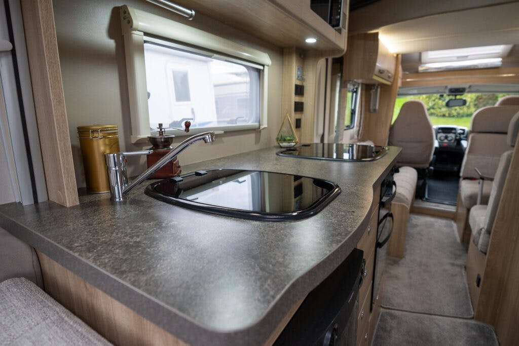 The interior of the 2019 Elddis Autoquest 196 Signature Edition motorhome showcases a kitchen area with a countertop. The countertop features a built-in sink with a faucet, a stove with a glass cover, and various kitchen items. In the background, there is a seating area and the driver's cabin.