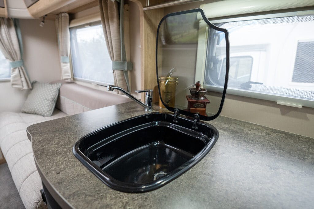 A 2019 Elddis Autoquest 196 Signature Edition campervan kitchen area features a black sink with a lifted glass cover. A modern faucet is installed beside the sink. In the background, there is a cushioned seating area, large windows with curtains, and a small countertop with a jar and decor item.