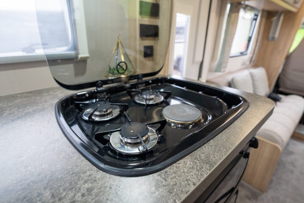A close-up view of a gas stovetop with four burners inside the 2019 Elddis Autoquest 196 Signature Edition's kitchen area. The stove is clean and has a glossy black finish. The countertop is grey with a textured surface. The background shows part of the camper's interior with seating and windows.