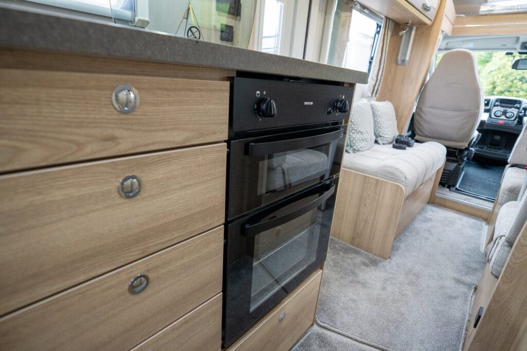 The image shows the interior of a 2019 Elddis Autoquest 196 Signature Edition motorhome, featuring a compact kitchen area with wooden cabinets and drawers. The kitchen includes a black built-in oven with two control knobs. The seating and driving area are visible in the background.