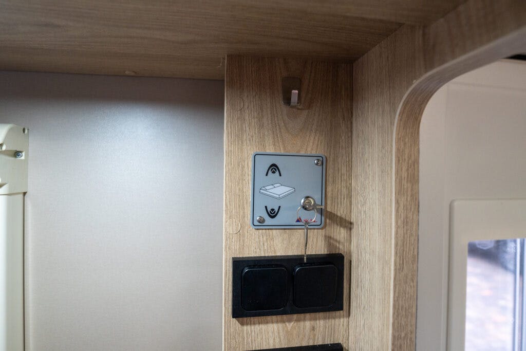 A close-up view of a wooden wall inside a 2019 Elddis Autoquest 196 Signature Edition camper, featuring a small control panel with a key inserted, and two black switches below it. The wall has a curved edge on the right and is adjacent to a white surface.