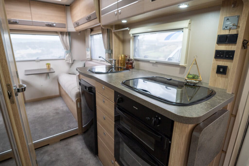Interior of the 2019 Elddis Autoquest 196 Signature Edition RV kitchen featuring wooden cabinetry, a dark countertop with a sink, a stove with a glass cover, and a black oven. A small window with curtains is above the sink. The adjacent sitting area has a window and a cushioned bench seat.