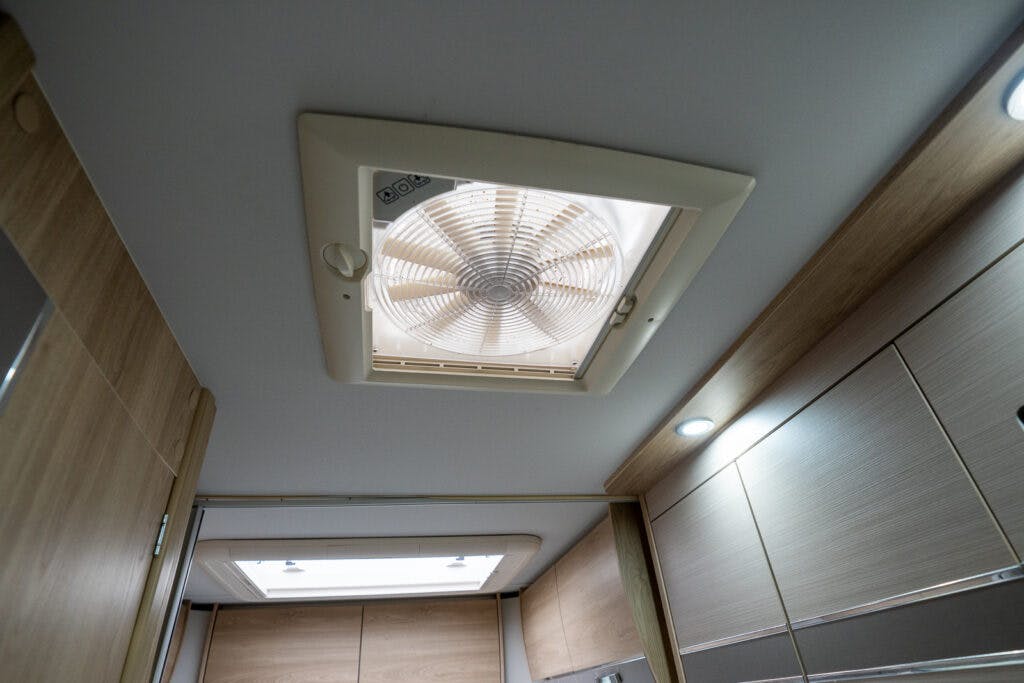 The image shows a ceiling vent fan installed in a well-lit wooden RV interior, specifically the 2019 Elddis Autoquest 196 Signature Edition. The vent features a circular fan protected by a plastic grid and is surrounded by a rectangular frame embedded in the ceiling panel. Cabinets and lighting fixtures are visible.
