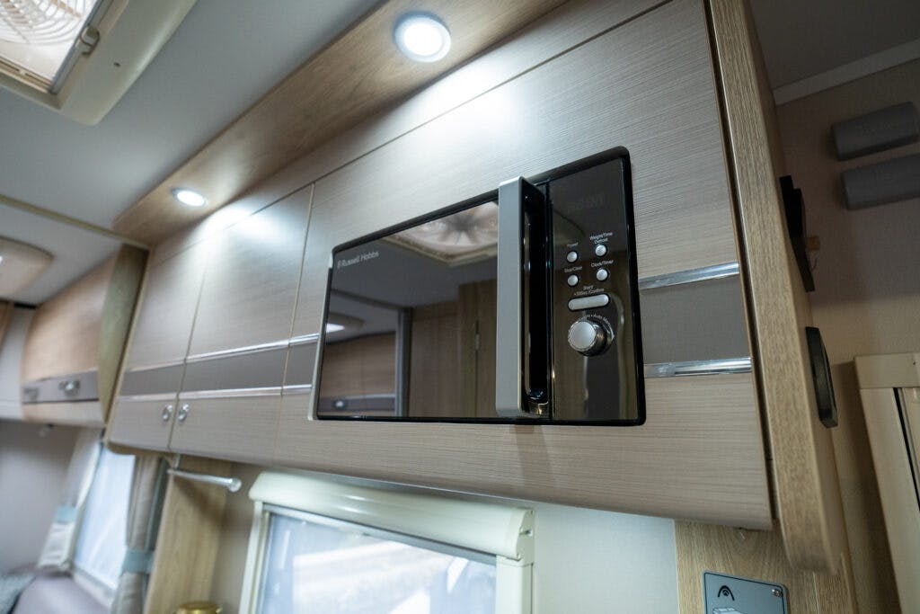 A built-in microwave oven is installed in a wooden cabinet with a light finish, just like the sleek design you'd find in the 2019 Elddis Autoquest 196 Signature Edition. The microwave has a black front with control buttons and a handle. Below the cabinet is a window with a rolled-up blind. The area is well-lit with ceiling lights.