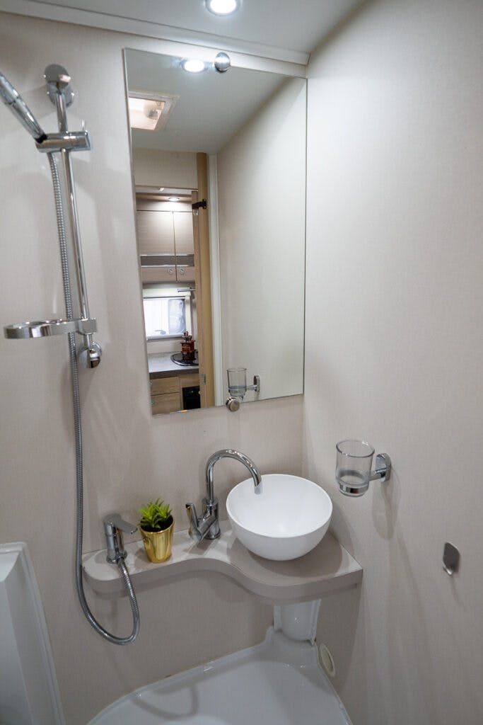 A compact bathroom in the 2019 Elddis Autoquest 196 Signature Edition features a white sink with a mounted faucet and a round mirror above it. Next to the sink is a small countertop with a potted plant. The shower area boasts a wall-mounted showerhead and hose, while light-colored walls keep the space well-lit.
