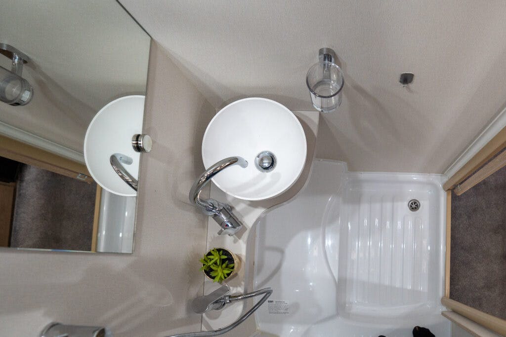 A compact bathroom in the 2019 Elddis Autoquest 196 Signature Edition features a white round sink and chrome faucet mounted on a corner counter. A mirror, toothbrush holder, small green plant, and a narrow shower tray with a drain are visible. The walls are light-colored, and the floor appears carpeted.