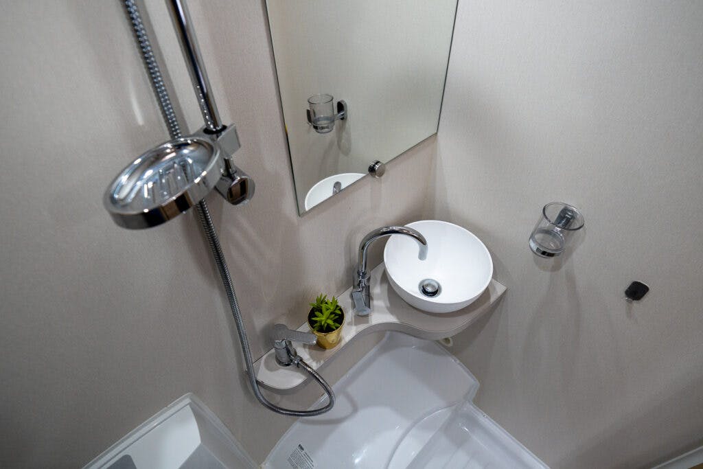 A compact bathroom inside the 2019 Elddis Autoquest 196 Signature Edition features a small round sink with a single-handle faucet, a wall-mounted mirror, a handheld showerhead on an adjustable rod, a small potted plant on the sink counter, and a corner bathtub. The walls are plain and light-colored.