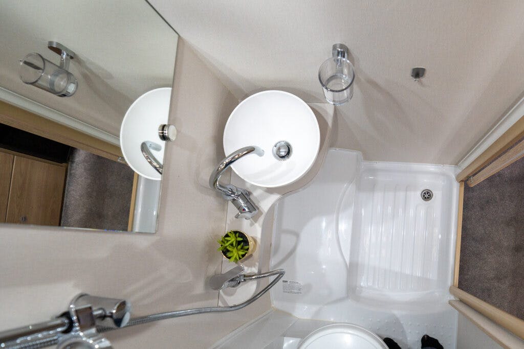 A compact bathroom in the 2019 Elddis Autoquest 196 Signature Edition features a white round sink with a chrome faucet, a clear soap holder mounted on the wall, a small plant on the counter, and a shower area with a white floor tray. A large mirror reflects these elements.