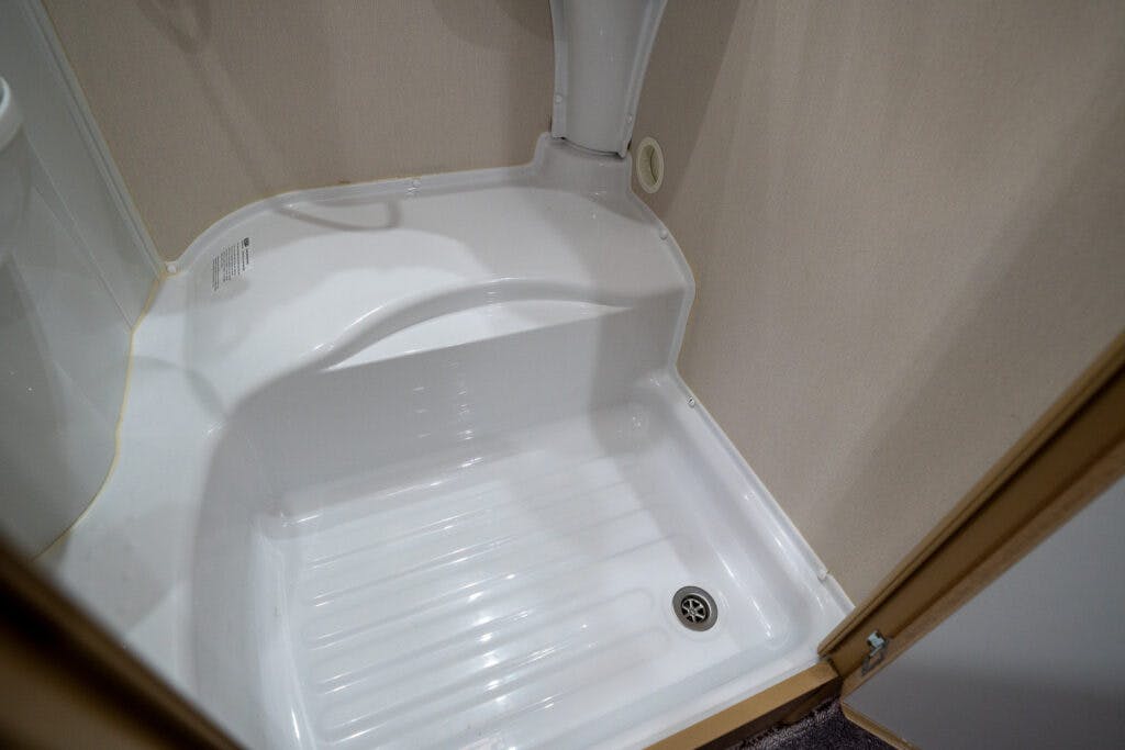 A close-up view of a small white corner shower with a raised seat in the 2019 Elddis Autoquest 196 Signature Edition. The shower features a textured floor for slip resistance and a metal drain cover. The beige walls and part of an adjacent carpeted area are also visible.
